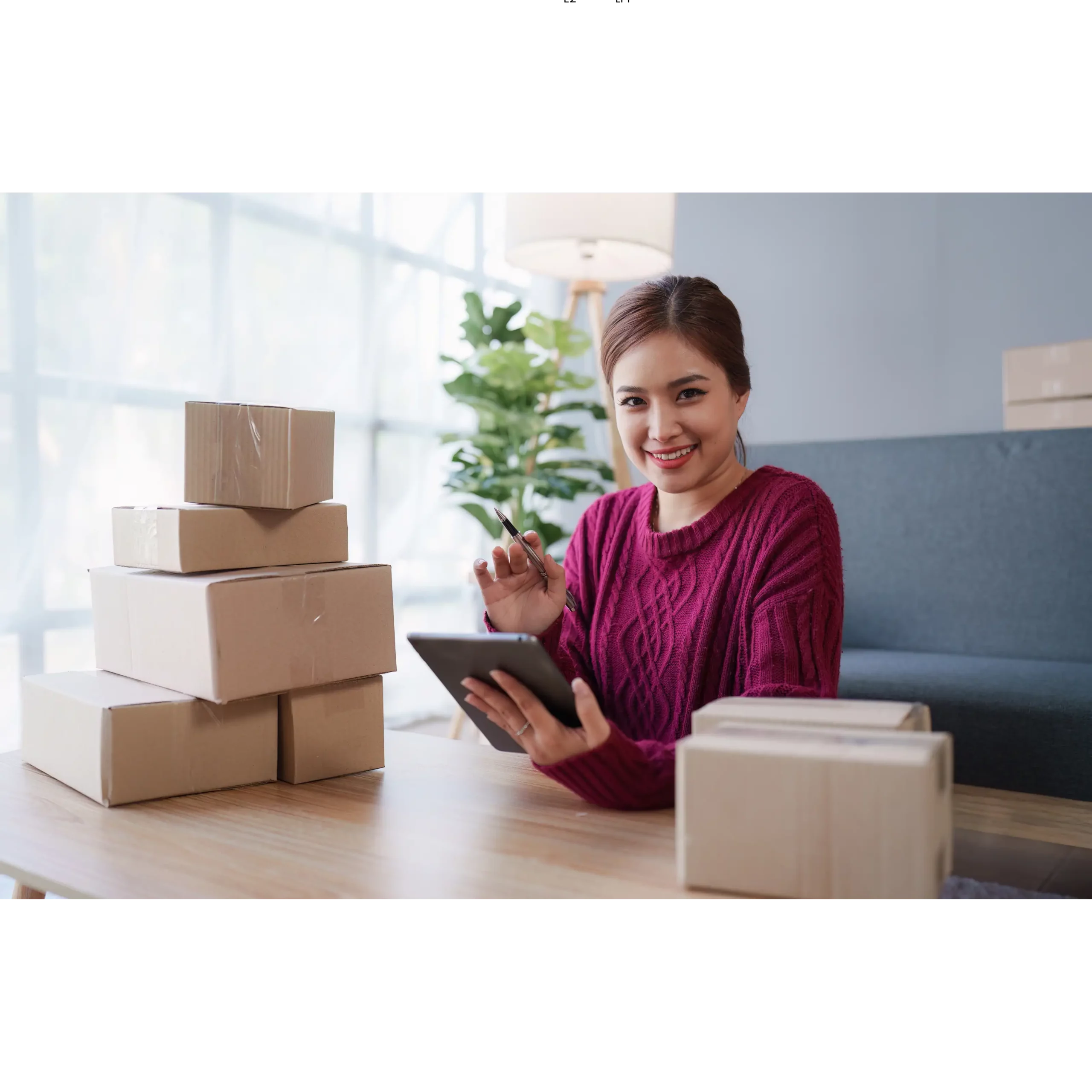 Femme asiatique travaillant sur une tablette, il y a des colis fermés sur la table autour d'elle - illustration d'une entrepreneuse PME prospère, représentant une solution RH pour les entreprises suisses.